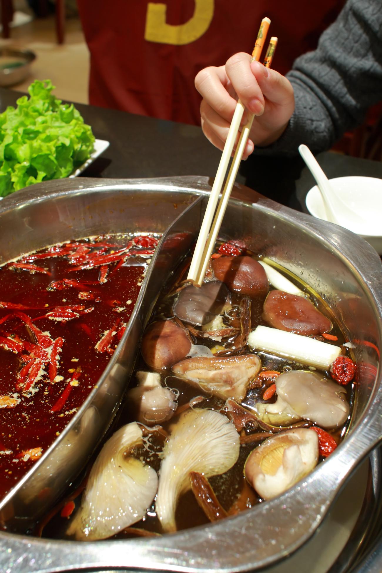 Photo of a Sichuan hot pot with a divider in the middle, and person holding chopsticks into the soup